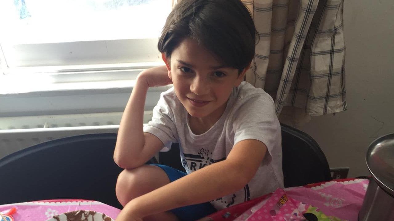 Oliver Archer as a young boy. He is sat on a chair, wearing a grey t-shirt and blue shorts. He has brown hair.