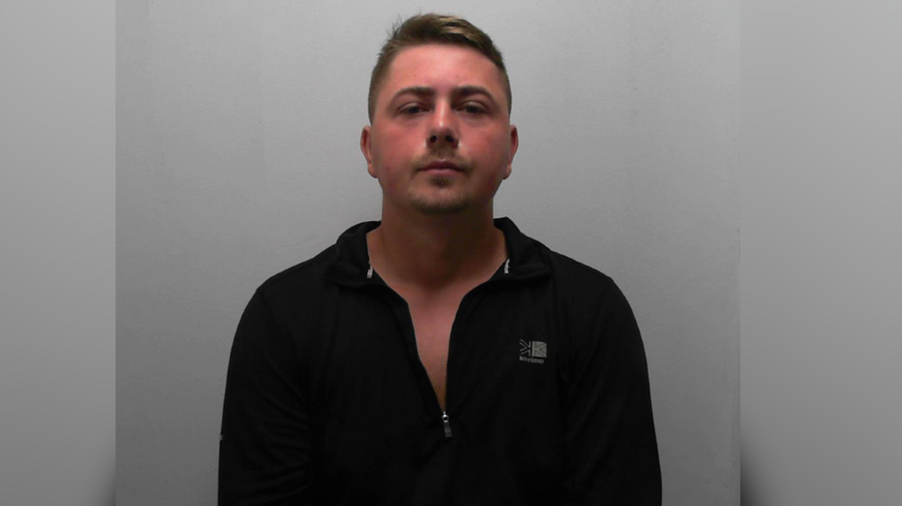 A police mugshot of a man wearing a dark open neck top with short hair staring at the camera