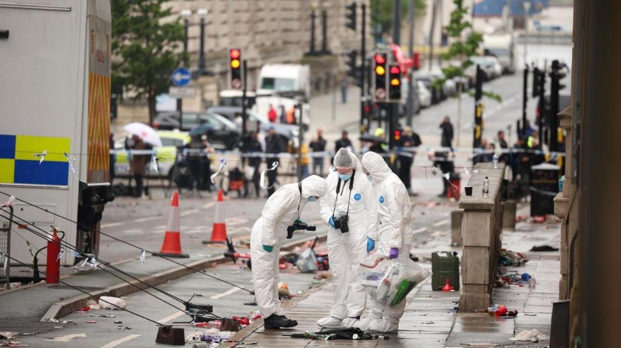 Three forensics officers, wearing white overalls and blue masks, are at the scene where a car collided with fans during the Liverpool FC trophy parade in Liverpool city centre.