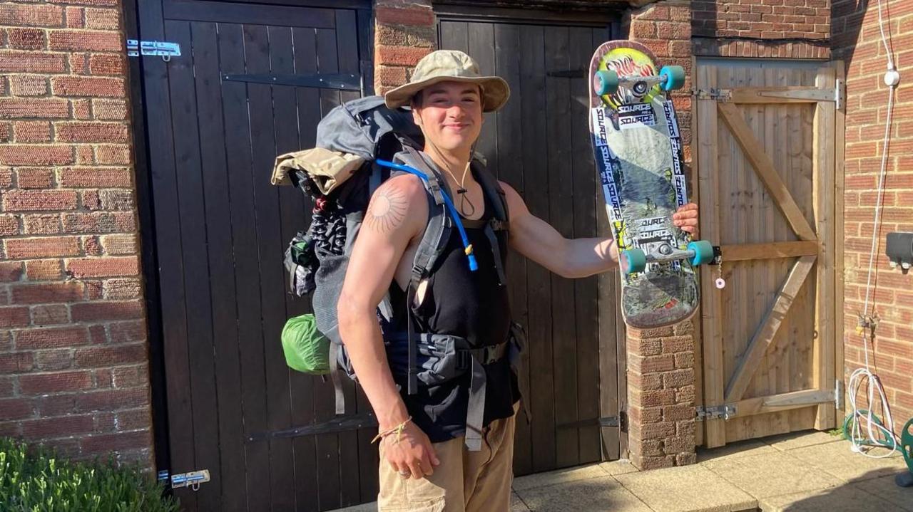 A young man with a hat and large backpack holds up a skateboard. 