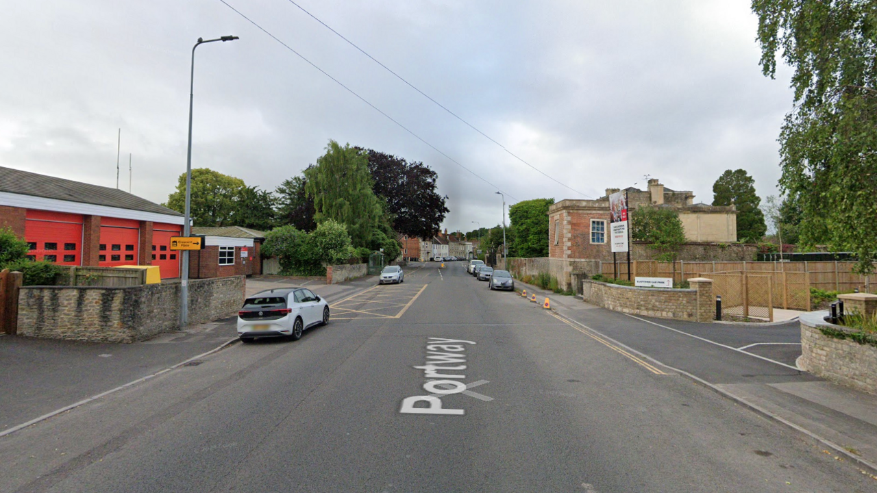 Portway Road in Warminster, Wiltshire. The fire station can be seen on the left. Other buildings can be seen on the opposite side of the road. Cars are parked up on either side of the road, next to the kerb. 