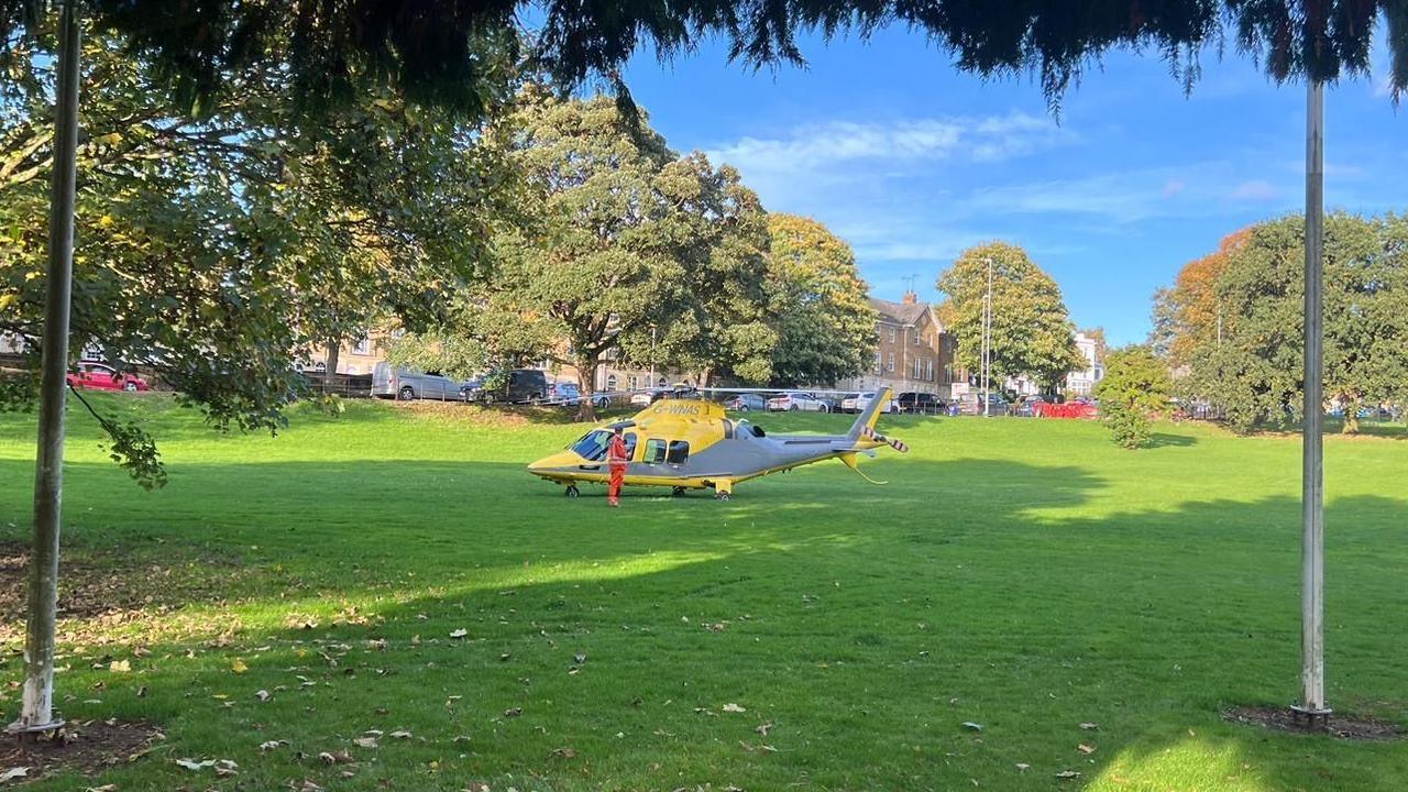 Large town centre park with trees in the background. A yellow air ambulance has landed in the centre of the grassed area and one person in orange overalls is standing outside it. A road lined with buildings is visible in the background.