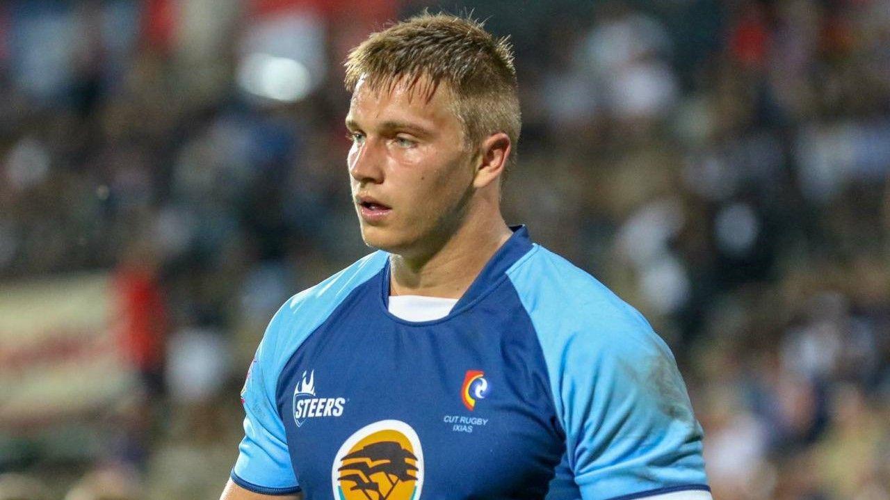 A young adult Rayno Nel seen from chest up looking tired during a rugby match. He wears kit with a royal blue chest, sky blue sleeves and a mustard sponsor's logo. A blurred crowd is in the background
