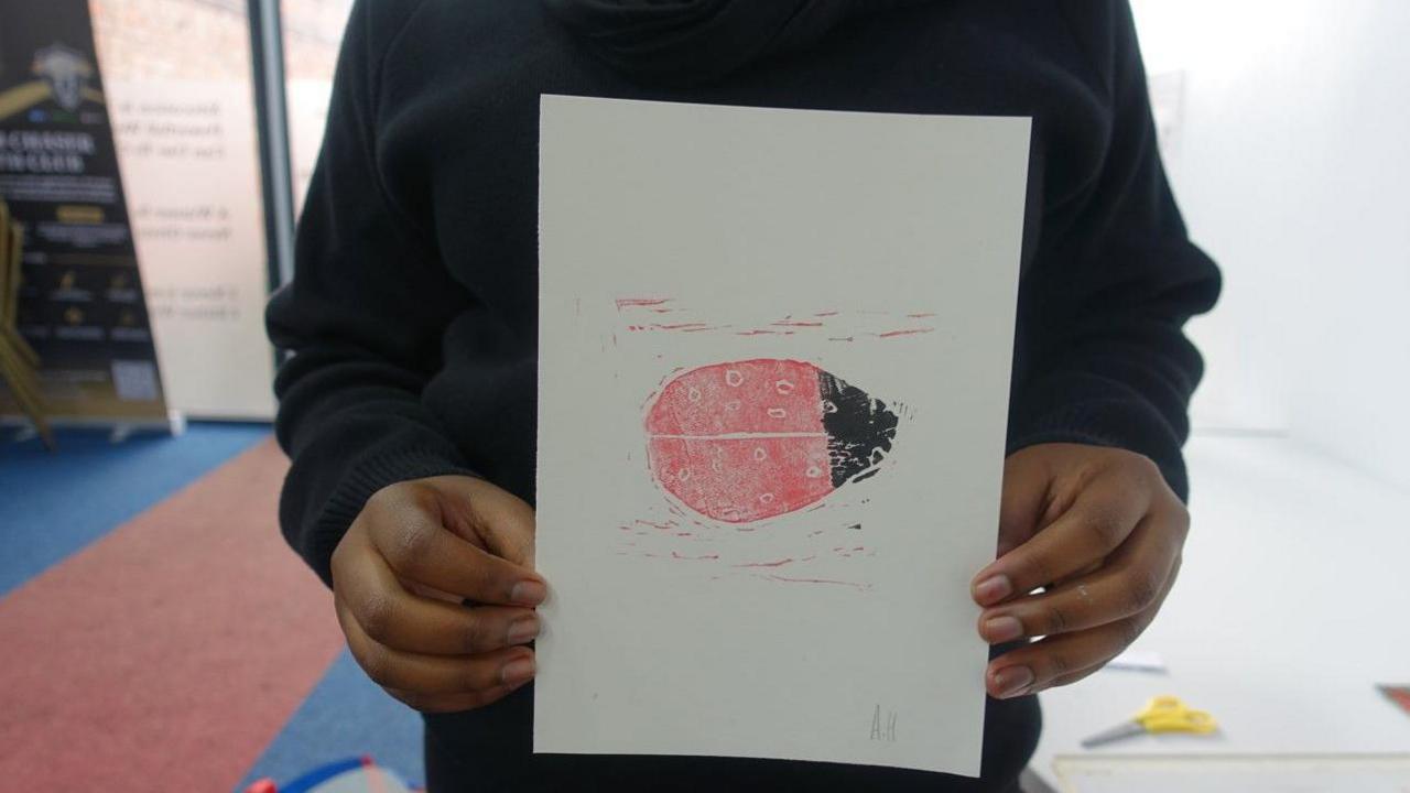 A child holds a picture of a red and black ladybird with the paper between their hands.