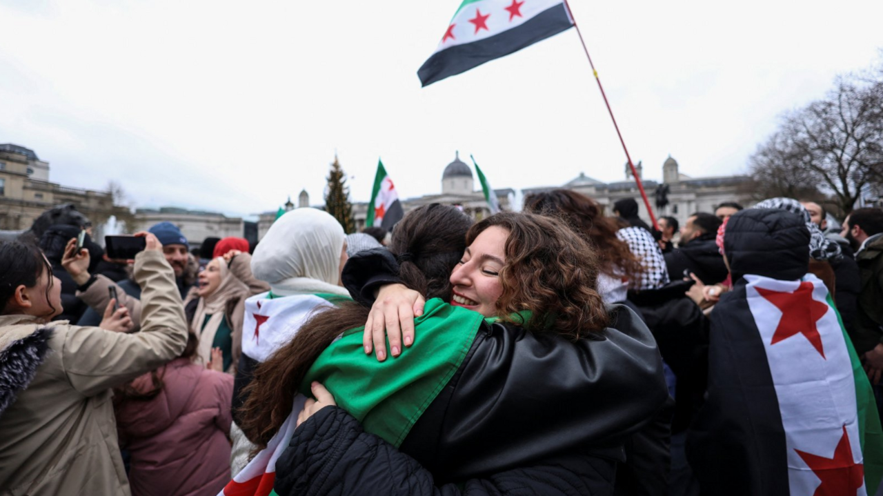 Two women - one wearing a Syria flag around her shoulders - embrace at a celebration to mark the end of Assad's regime at London's Trafalgar Square on an overcast December day 