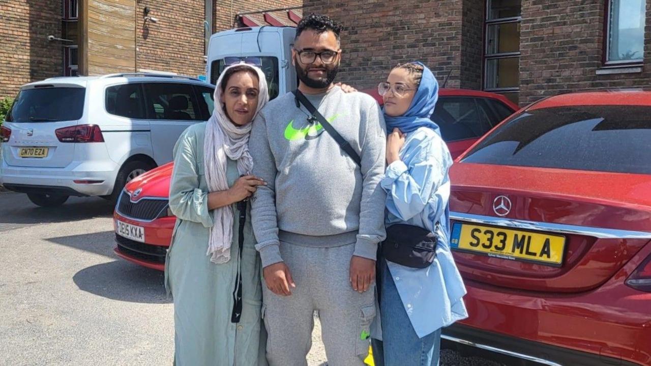 Jamil Hussain standing with his mum and sister and posing for a photo. He is wearing a full grey Nike tracksuit and a black cross body bag. His mother is wearing a light blue abaya dress with a white headscarf. His sister has braided hair and is wearing blue jeans, a blue oversized shirt and a blue headscarf. They are standing in a car park on a sunny day.