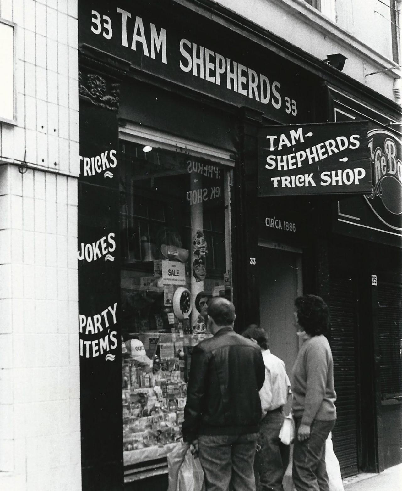 Exterior of Tam Shepherds - Shop front painted black with white text reading "Tam Shepherds" with an awning sign reading "Tam Shepherds Trick Shop