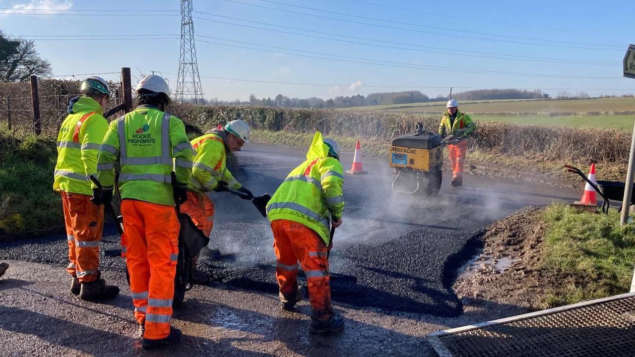 A group of workmen in fluorescent clothing on a roadside work to lay down tarmac. 