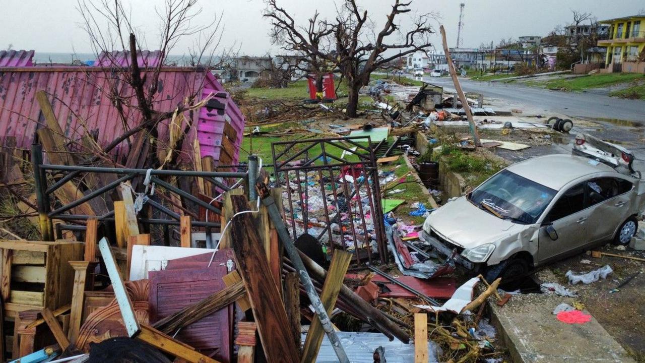 A house which has been destroyed in the hurricane, plus a car which has been damaged, and a road littered with debris. 