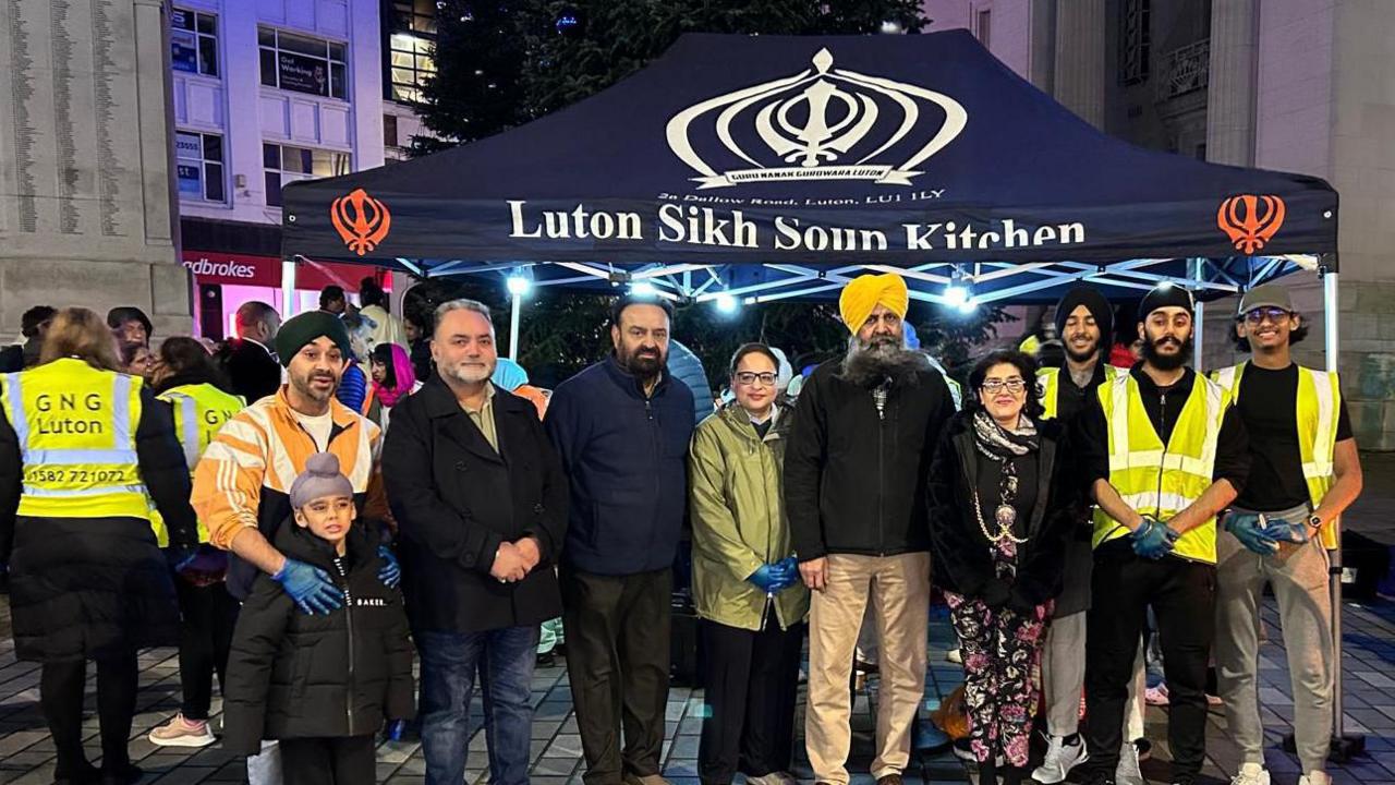 The gazebo for the Luton Sikh Soup Kitchen, which is navy blue and has a sikh symbol on that looks like a turban-shaped crown. In front of it stands a group of volunteers who are mostly men. Four of them wear turbans as well as a younger boy. Next to them are volunteers with their back turned who wear high vis jackets saying GNG Luton.