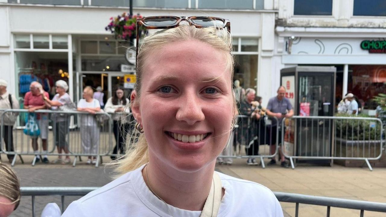 The photo shows a woman smiling at the camera. She has blonde hair which is tied up into a pony tail, and has sunglasses on her head. She's wearing a white t-shirt. She's standing on a high street, and there are various shops and people behind her. 