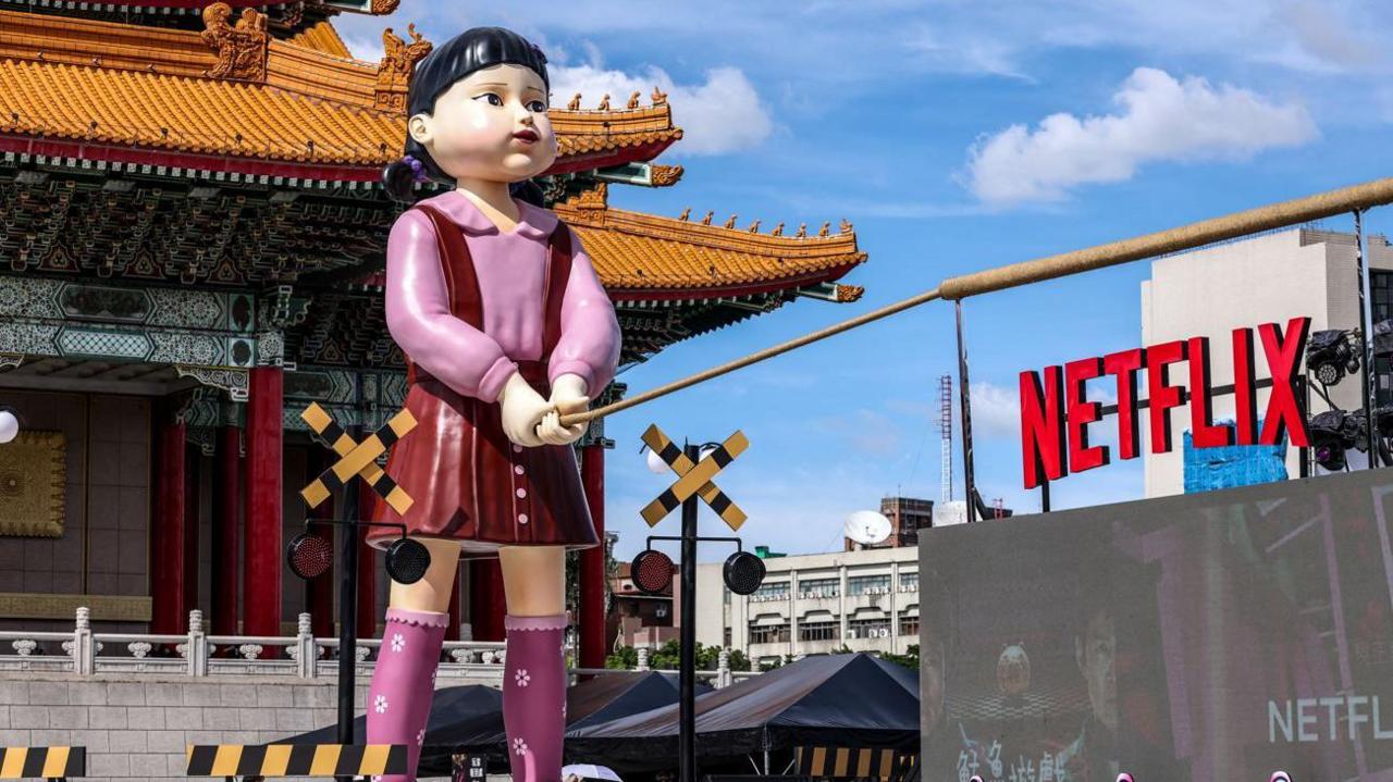 A giant sculpture of a Korean doll - a girl with black hair with a fringe and pigtails, wearing a red dress and pink blouse from the show Squid Game stands in front of a Korean temple, holding what appears to be a tug-o-war rope, next to a sign of the word Netflix.