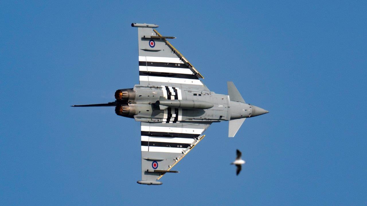 A grey military plane flies against a deep blue sky. 