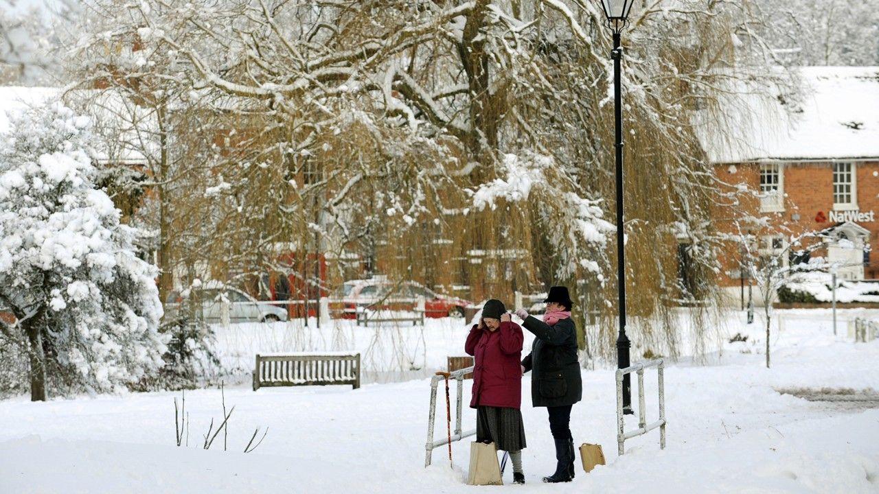 Two women in the snow adjusting a hat with a house and parked cars behind snow-filled trees