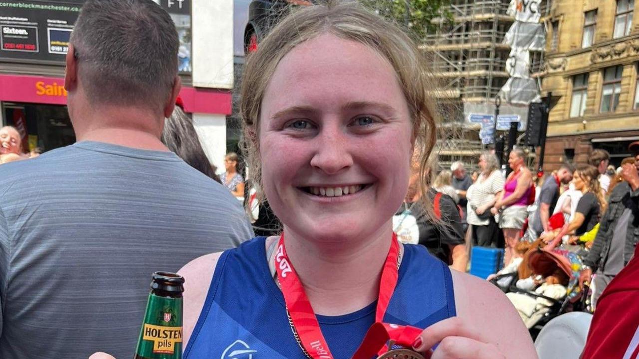 Isla Bayliss is smiling facing the camera. She is holding a bottle of lager in one hand and a medal in the other.