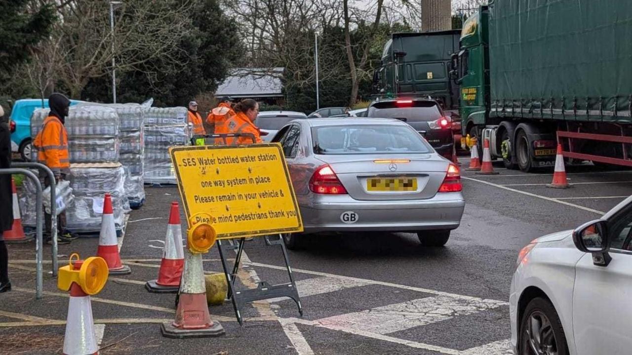 A yellow sign on the side of the road which says "SES Water bottled water system, one way system in place." It asks drivers to stay in their vehicles and mind pedestrians. A queue of cars is seeing passing workers in hi-viz clothing and pallets of bottled water.