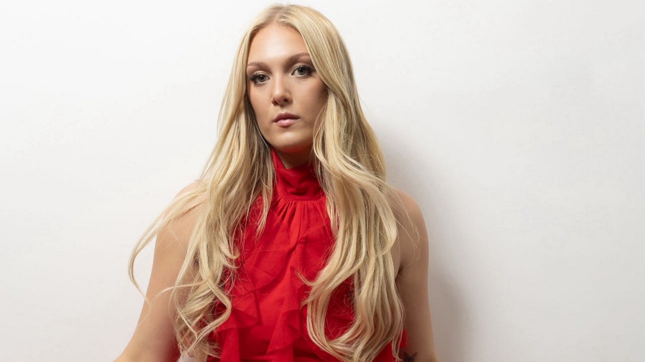 Emily Fern is posing for the camera in front of a white wall. She has long, wavy, blonde hair and is wearing a shoulder-less chiffon-style red dress with a ruffled bodice. She looks serious.