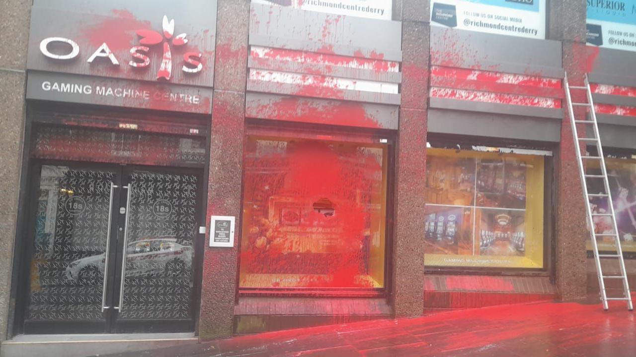 A shop front in Londonderry city centre partly covered in red paint following a vandalism attack. There are three large windows and a glass door all splattered with paint that is now partly cleaned. A ladder can be seen up against the building.