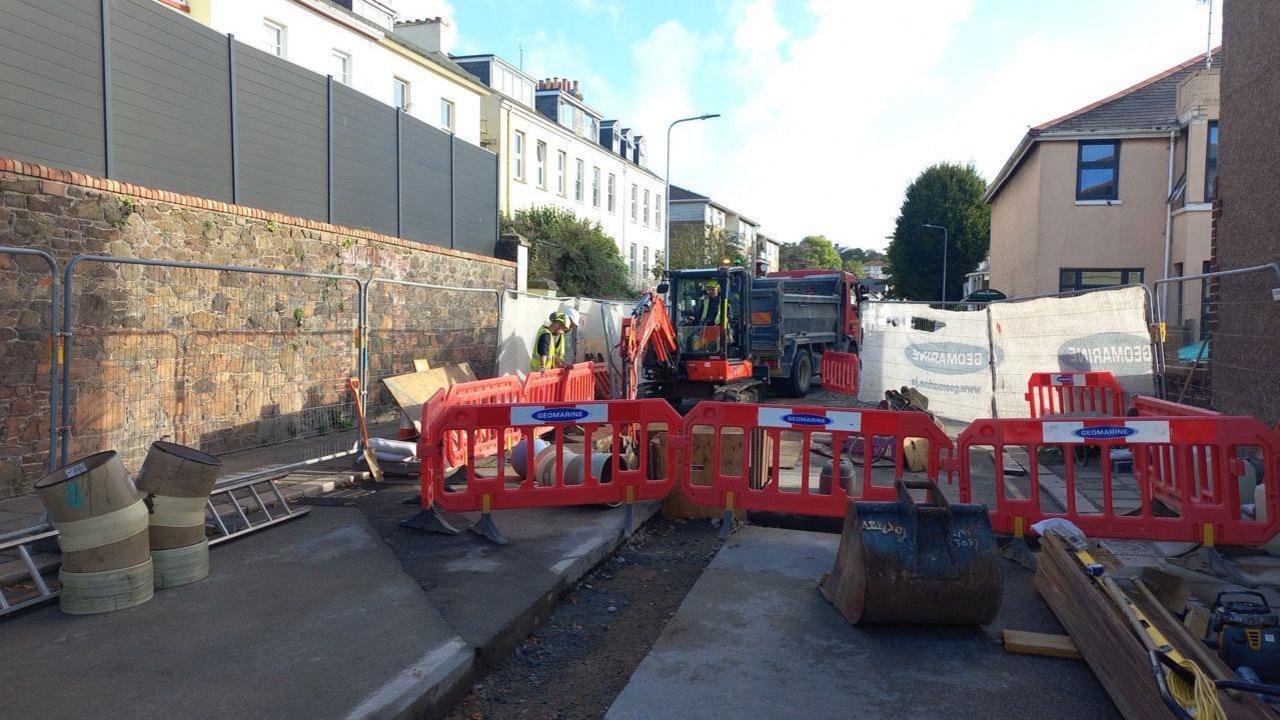 A road with temporary metal fencing around it. There are workers in high vis. There is a digger. There are houses around the road. The sky is blue with white clouds. 