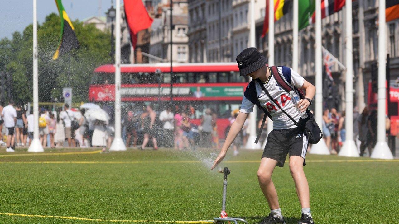a young person with a hat on and a water bottle in their hand puts their hand over a sprinkler in an attempt to keep cool, with white buildings, international flags on high poles in background, dozens of people and a red double decker bus