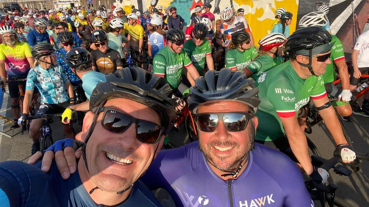 Graham smiles as he takes a selfie with a friend and they're at the start of a bike race with numerous other participants in the background, they're wearing helmets and sunglasses