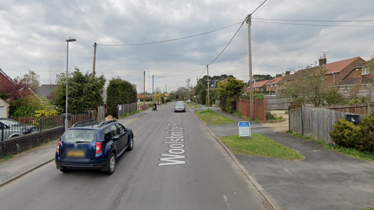 A Google street view screenshot of a residential road with semi-detached houses on both sides, with large font gardens. There are two car and a motorcycle driving down the road and it's a cloudy day.