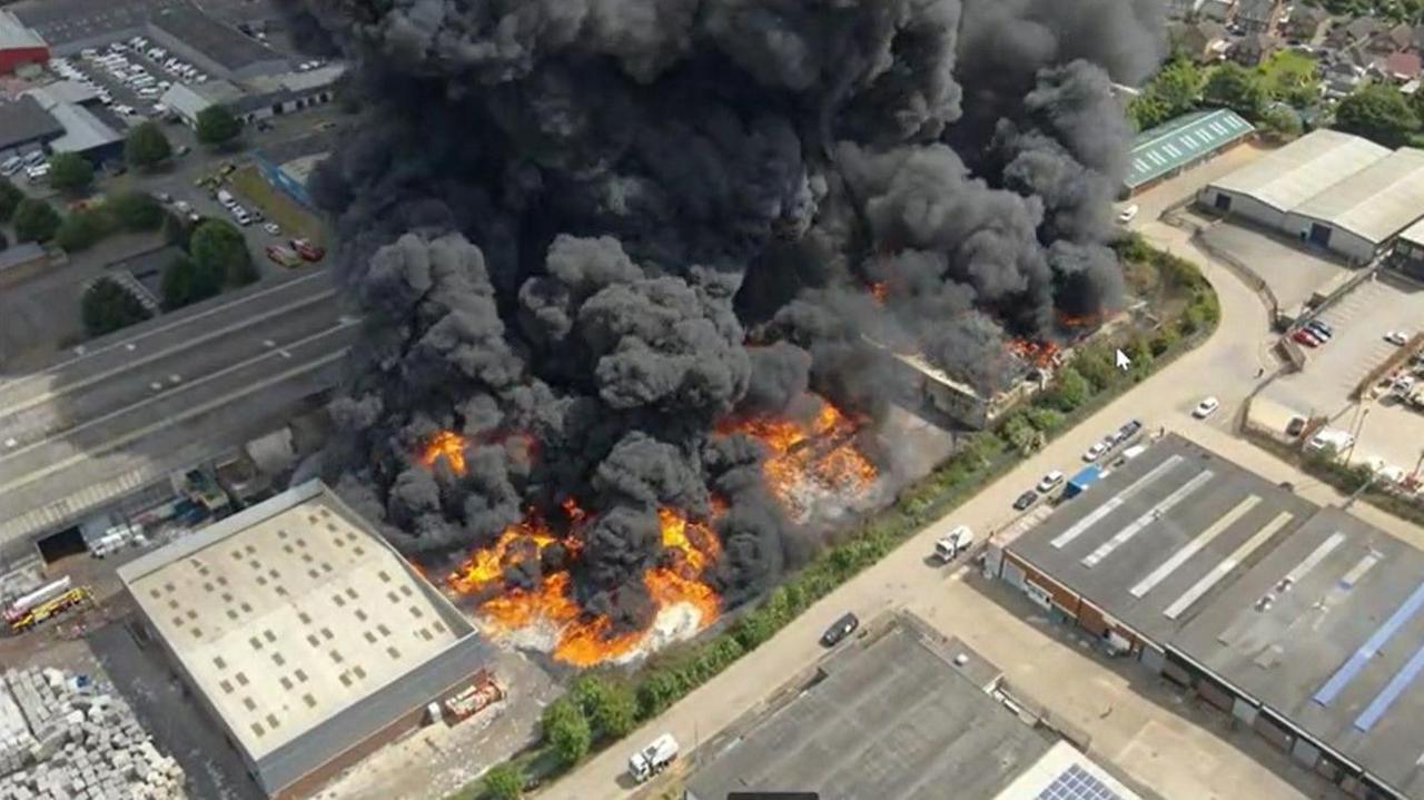 An aerial shot of a large recycling warehouse fire, showing flames and large amounts of smoke, buildings around it and cars on the road.