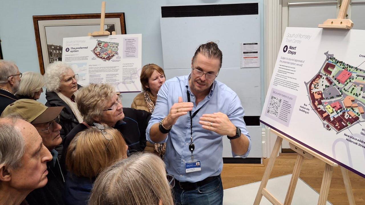 A small crowd listens to a planning officer speaking in front of a map of the proposed development on an easel.
