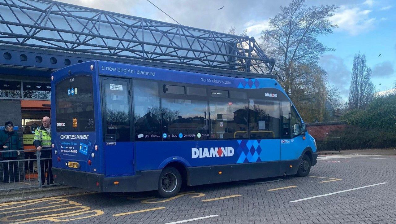 A blue bus with the word Diamond is parked in front of a bus station with a blue metal structure for a roof. It is a single decker bus in this generic image.
