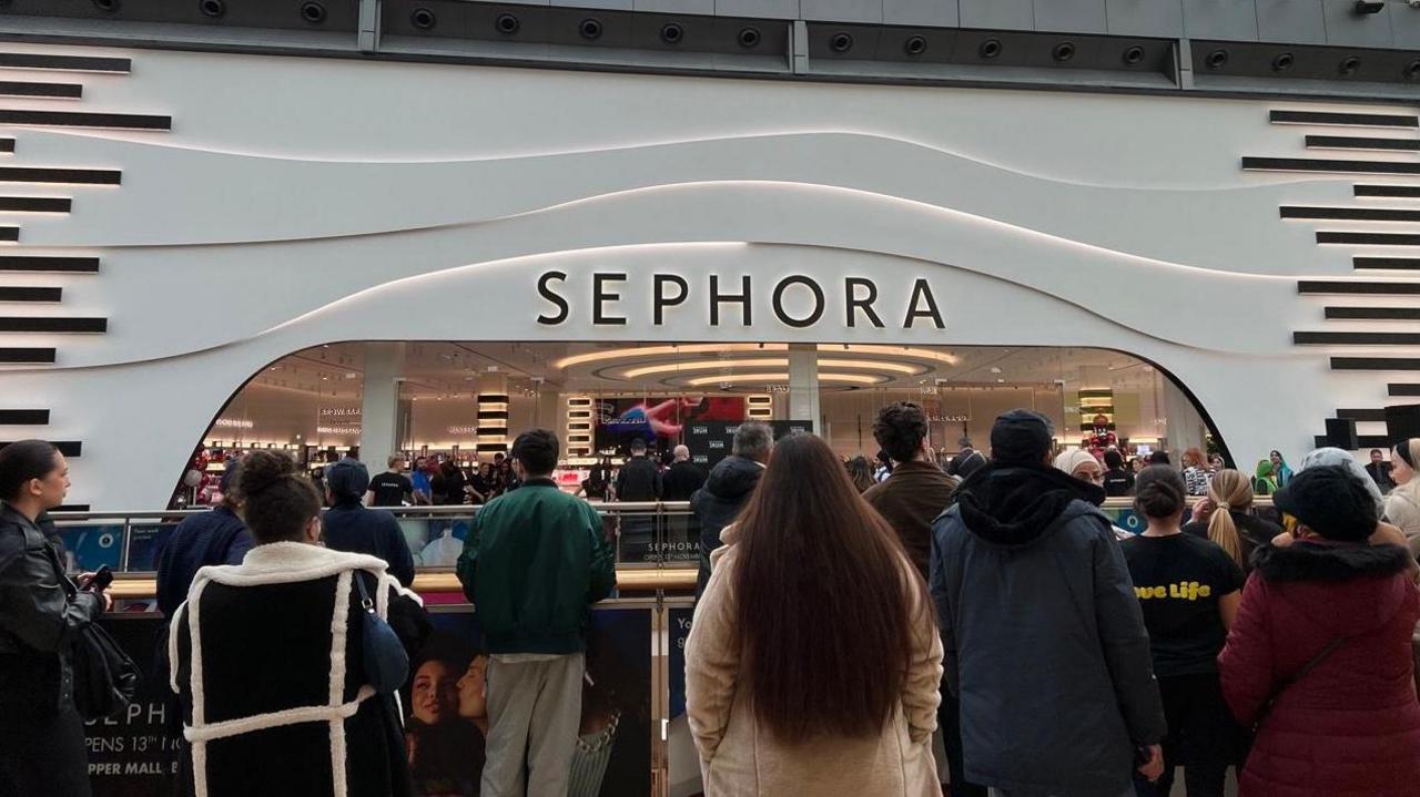 A large black and white fronted building with a wide entrance and a sign that says Sephora in black capital letters along the top. In the foreground shoppers can be seen facing the entrance to the shop.