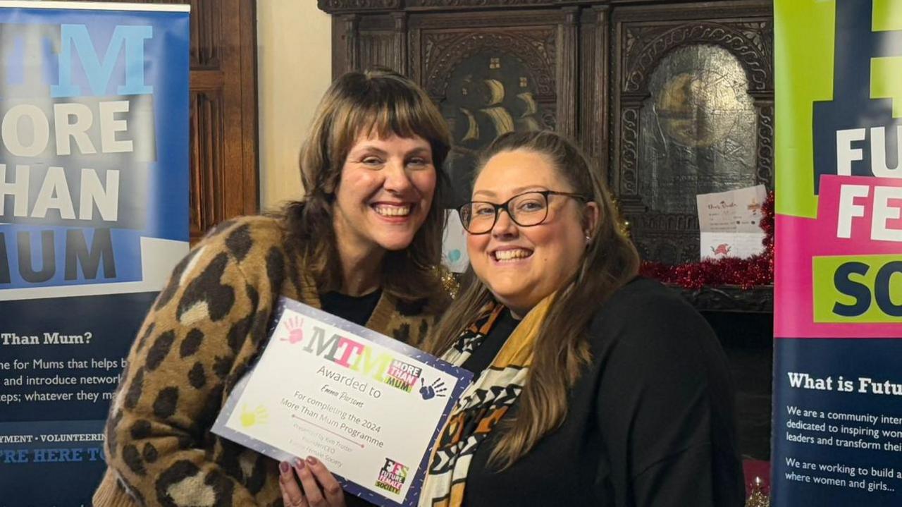 Kim Trotter and Emma Parson stand side by side smiling at the camera. Ms Trotter has shoulder length brown hair and is wearing a leopard print coat. Ms Parson has long brown hair and is holding a certificate. 