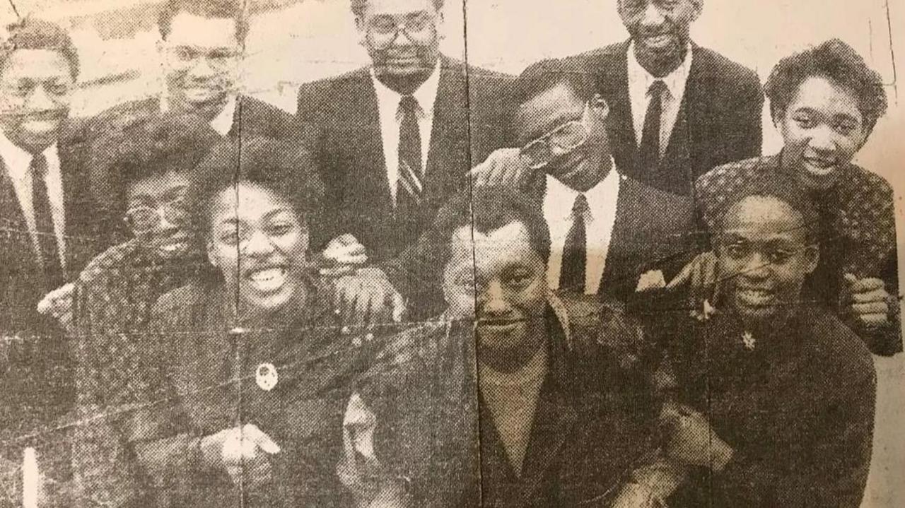 A newspaper cutting shows the singer with choir members in a black and white photo, The singer is stood in the middle with choristers standing around him holding his arms and smiling.