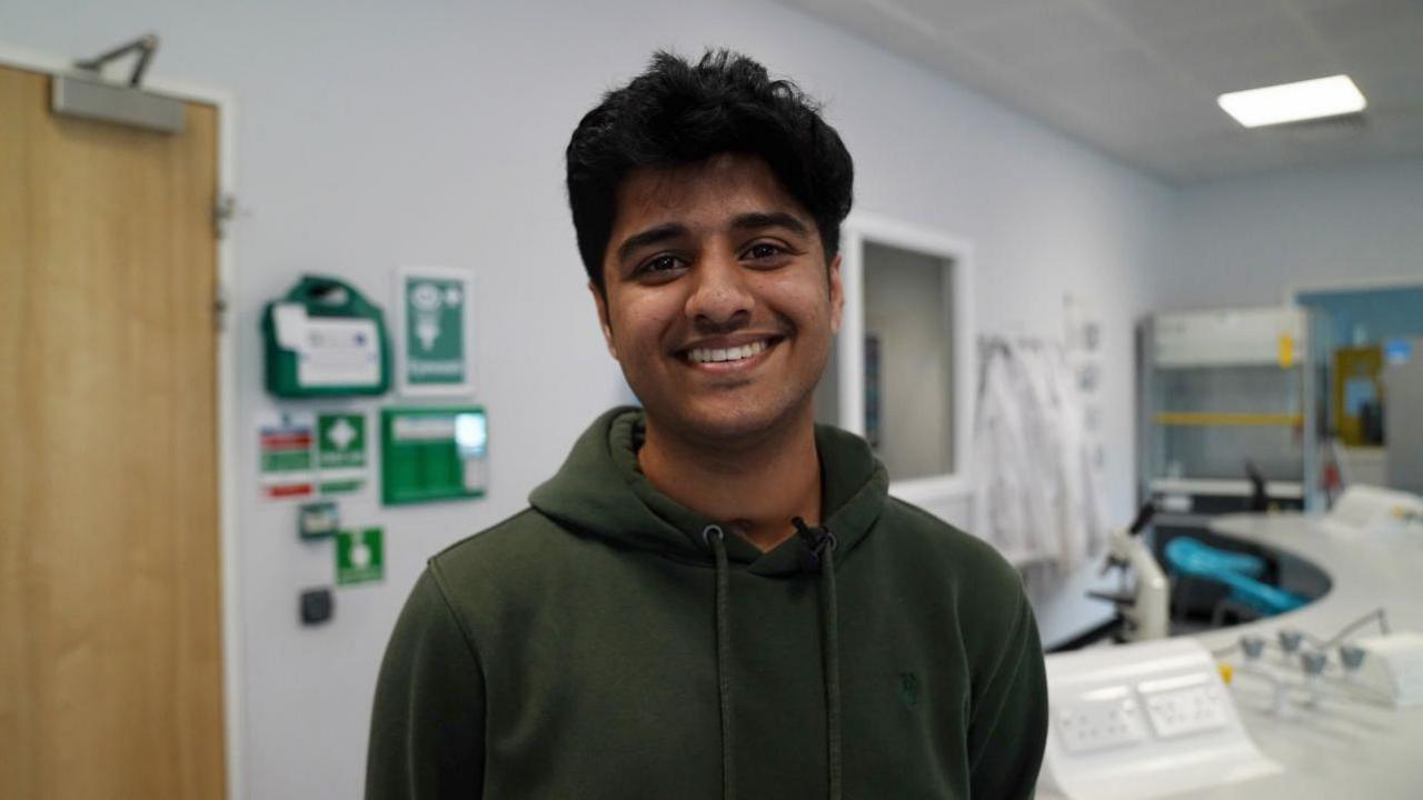 Eshan stands in a classroom and smiles at the camera. He has short, black hair and is wearing a green hoodie. 