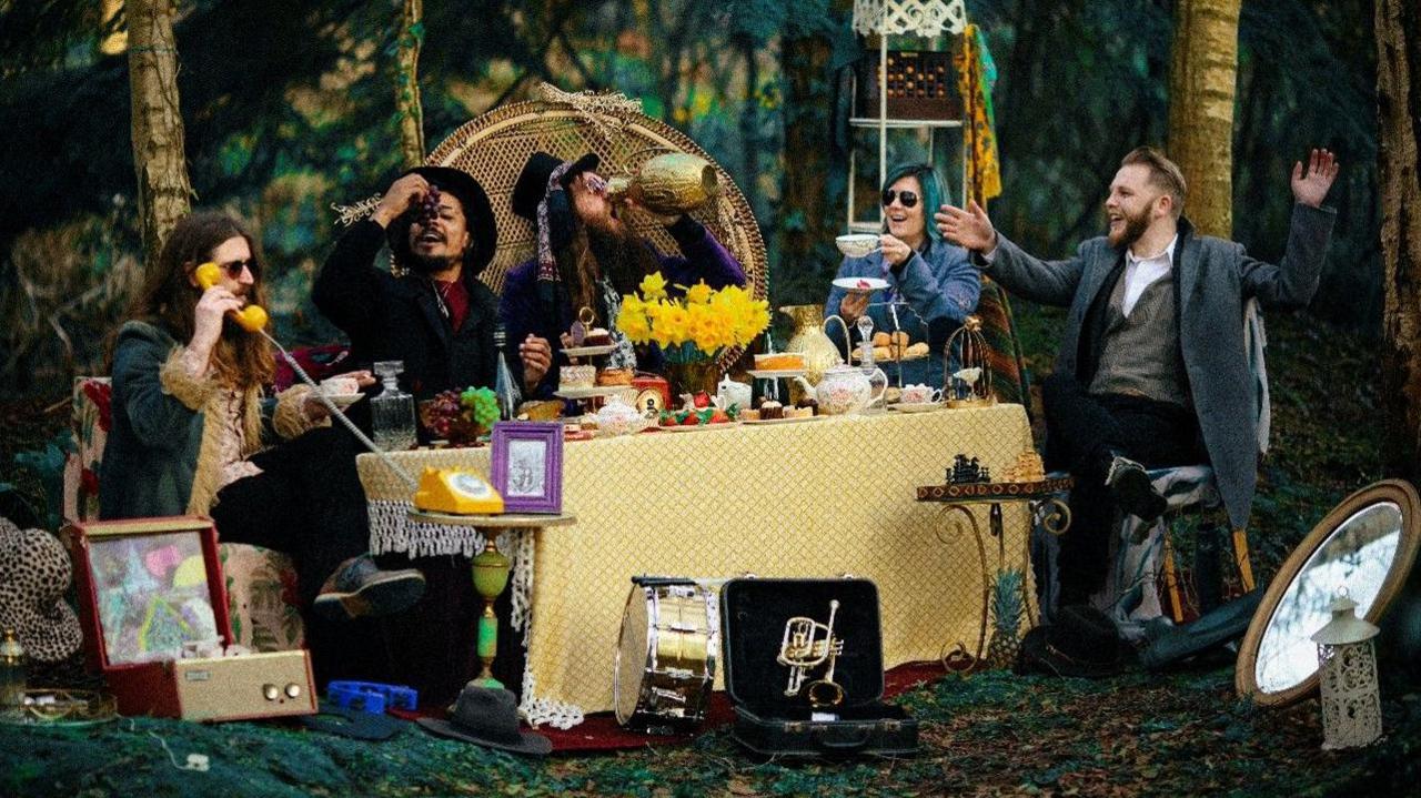Five members of a band sitting around a table covered in a yellow cloth. They are eating an afternoon tea while Richey is talking on a yellow phone. One member of the band is swigging out of a brass jug and another is eating a bunch of grapes.