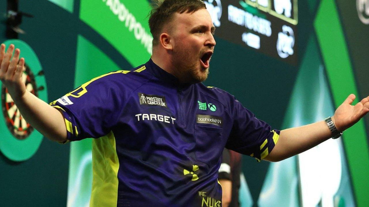 Luke Littler celebrates with his arms and mouth open. Part of a dart board is blurred in the background. 