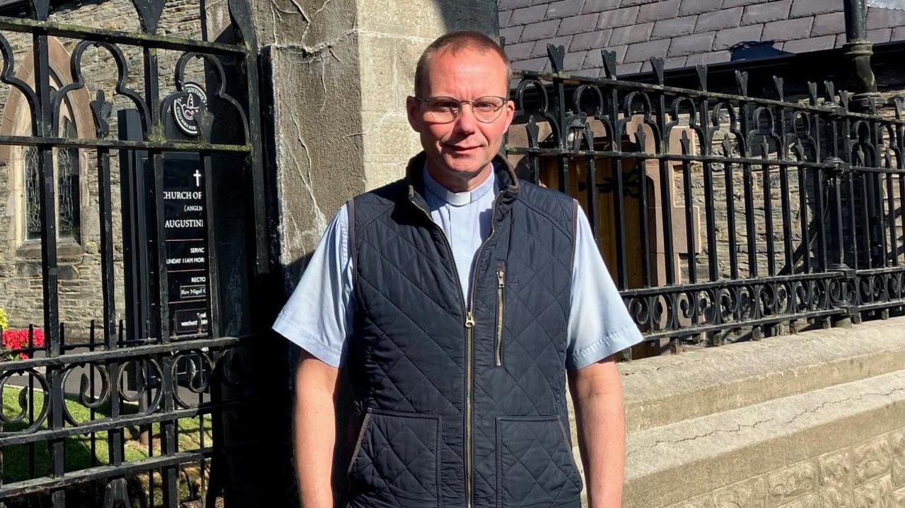 An Anglican rector stands in front of an old church. he is wearing a blue shirt and darker blue jacket. the church building can be seen behind him through iron railings
