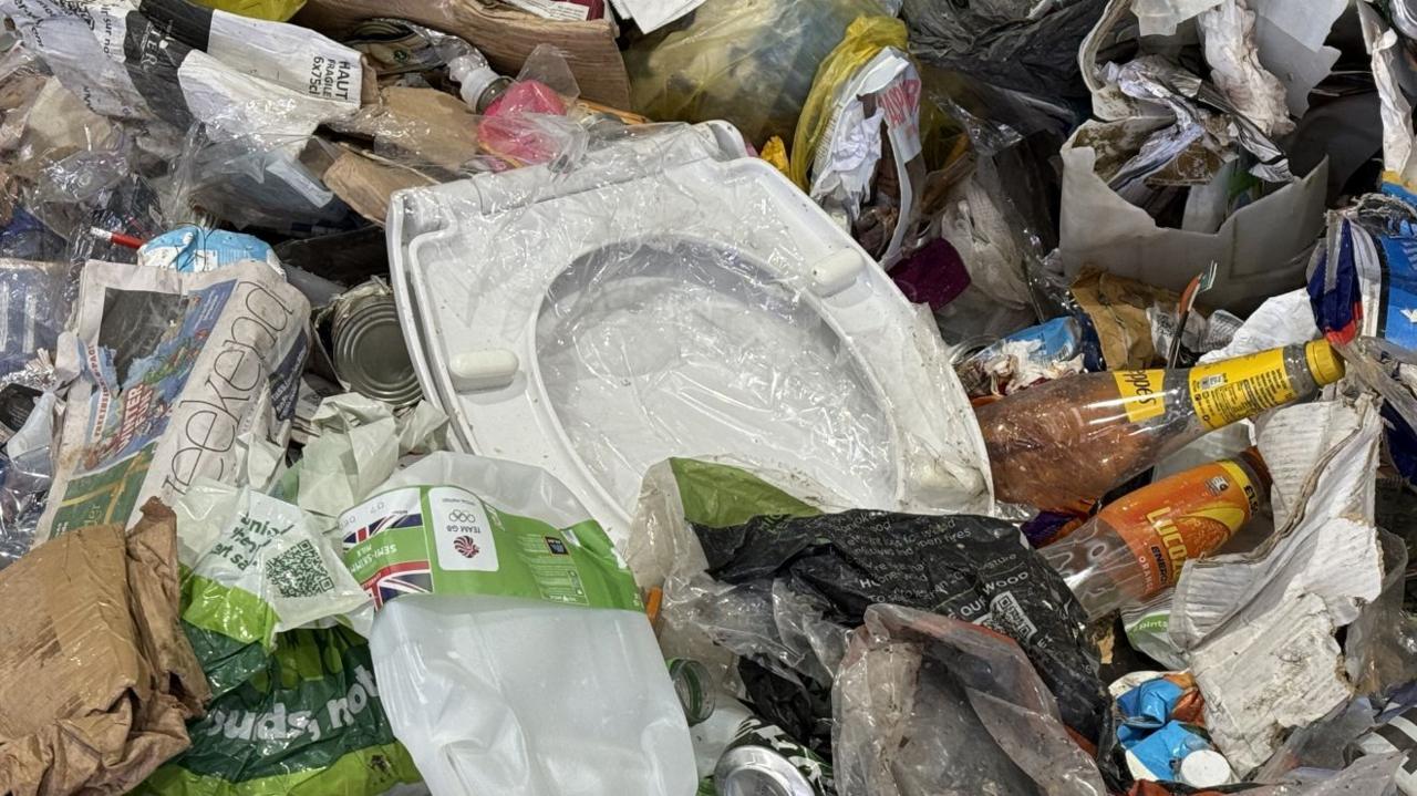 A white toilet seat, wrapped in plastic, sits among a pile of waste including glass and plastic bottles, newspapers, cardboard and plastic bags.