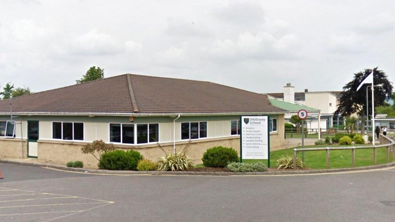 A screenshot from Google street view showing the one storey building at the entrance to Wellsway School. It is a low rectangular building with windows, a tiled roof and a grassy area to the right with bushes planted in a flowerbed. There is a sign for the school and a white flag pole.