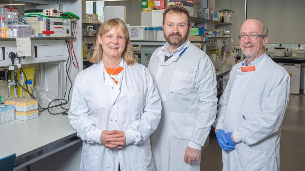 A woman and two men wearing white lab coats stand smiling in front of laboratory equipment.