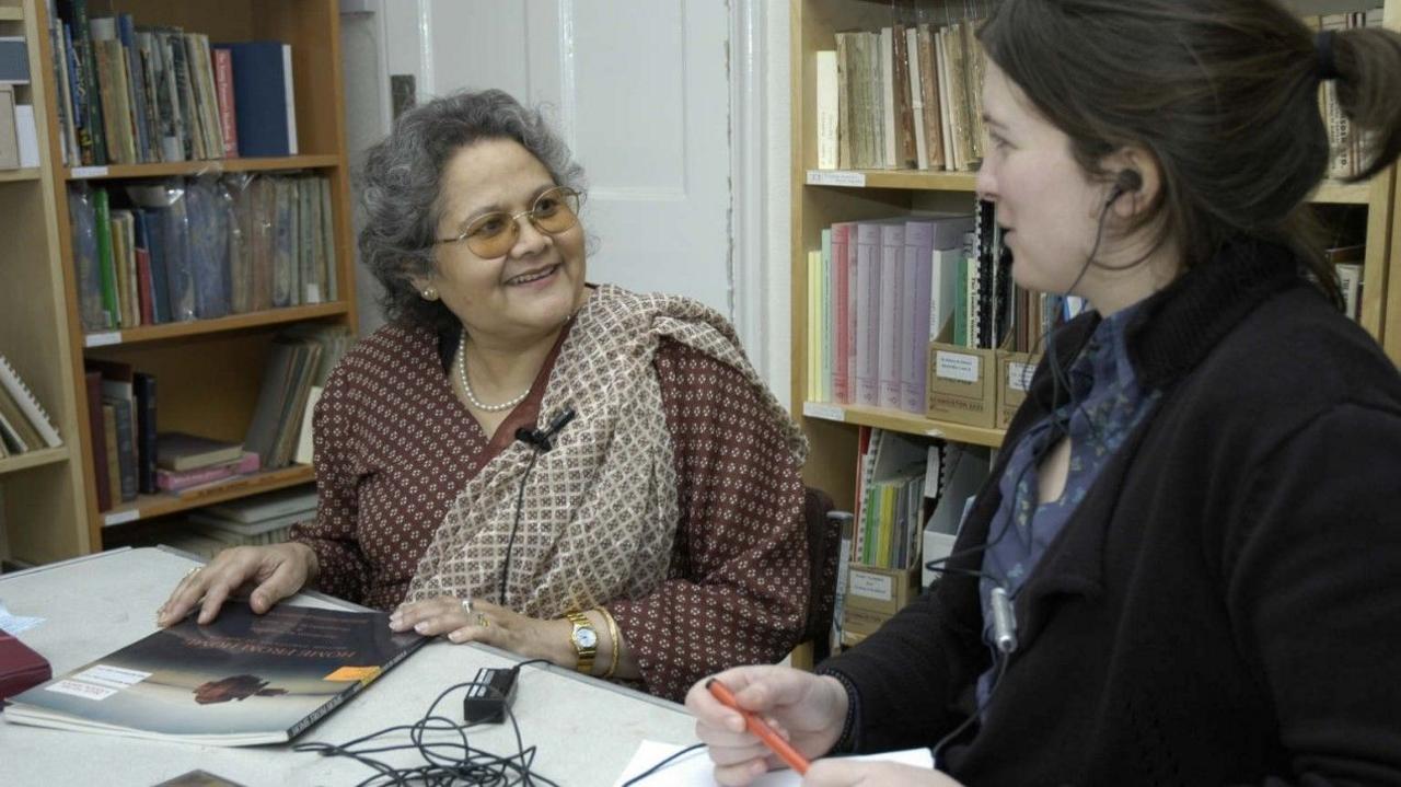 Two women in a room with folders and behind them on shelves. One woman, with grey hair and glasses is looking at another woman, smiling, while the other woman, on the right is listening to what she is saying, recording her as part of an oral history project. 