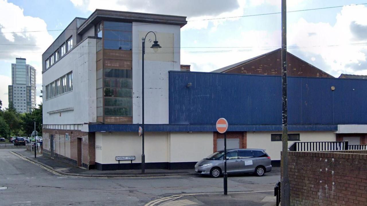 A old building that looks empty. It has a white exterior and a blue back end. There is an uber parked outside near a stop side. 