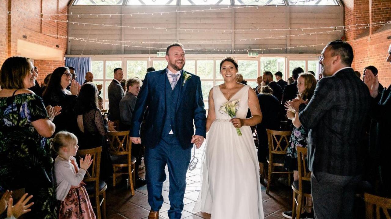 A man in a blue suit is holding hands with a woman in a white wedding dress as people on both sides applaud.
