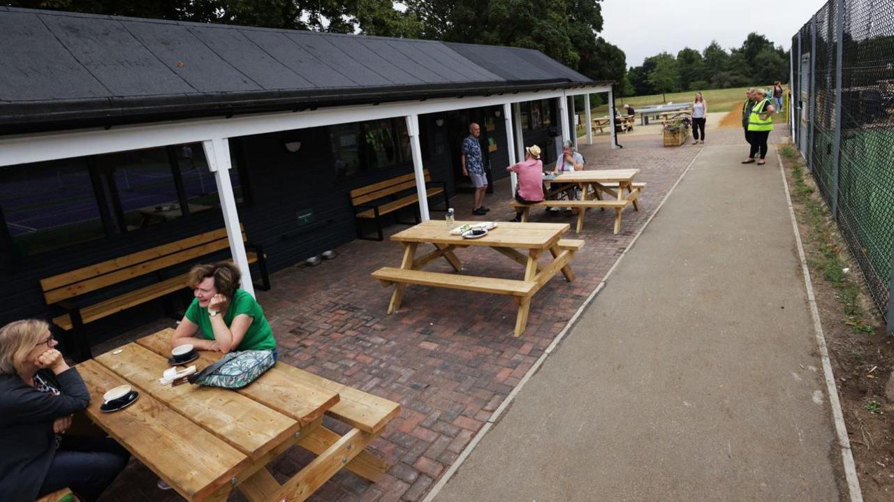 People sitting at or standing around outdoor tables at a café opposite a tennis court. It is an overcast day.