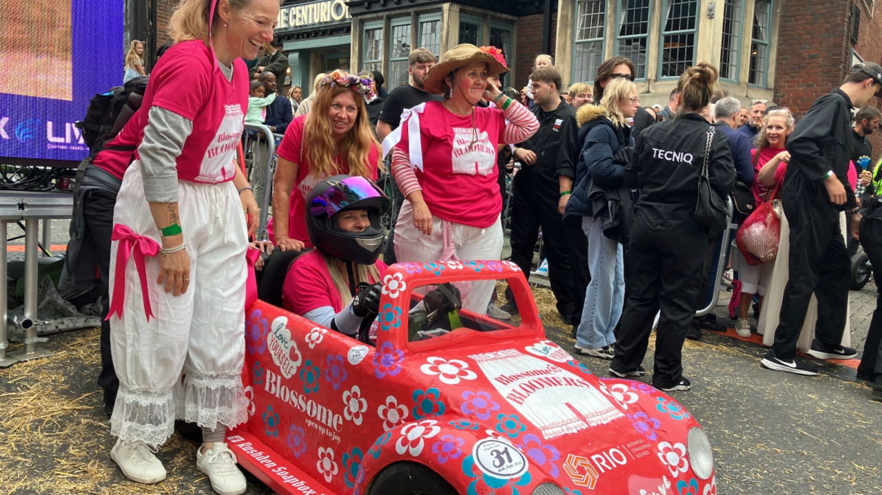 A woman is sat in a small red car-shaped kart. It kart is painted red and has flowers and a sign on the side which says Blossome. Three smiling and laughing women are stood behind the kart wearing a pink top and white trousers.