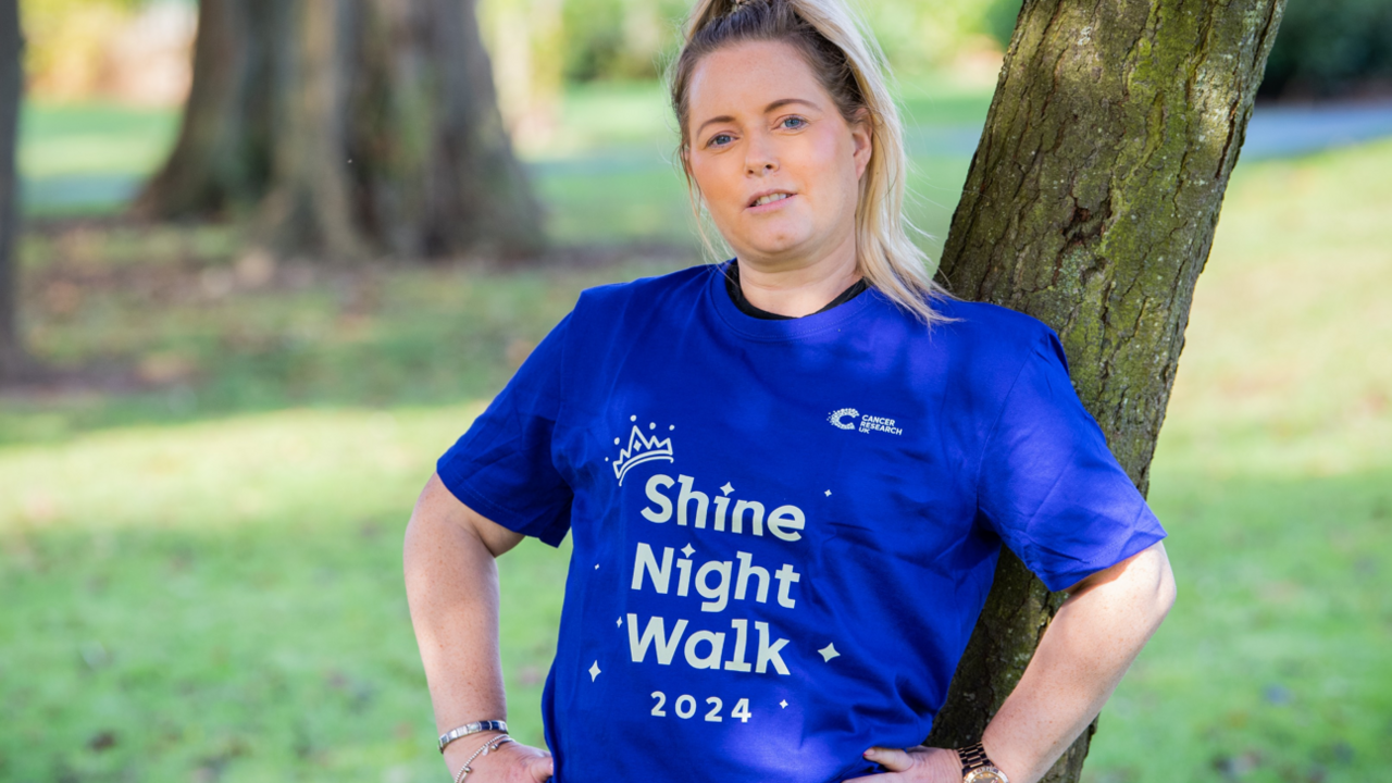 Stacey, wearing a royal blue Cancer Research UK t-shirt, stands outside with her back to a tree 