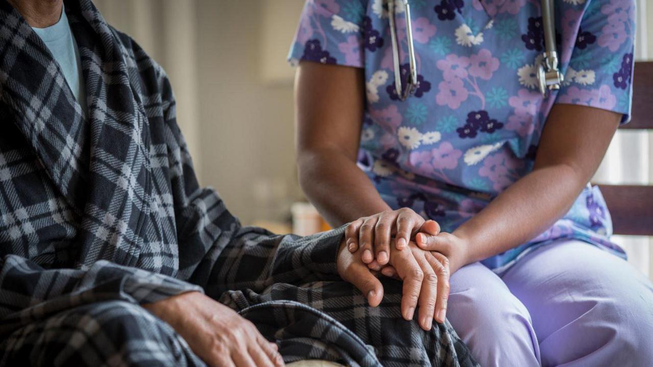 A stock image of a man in a black and white tartan dressing gown sat down next to a nurse in a purple outfit with a medical device around her neck holding his hand