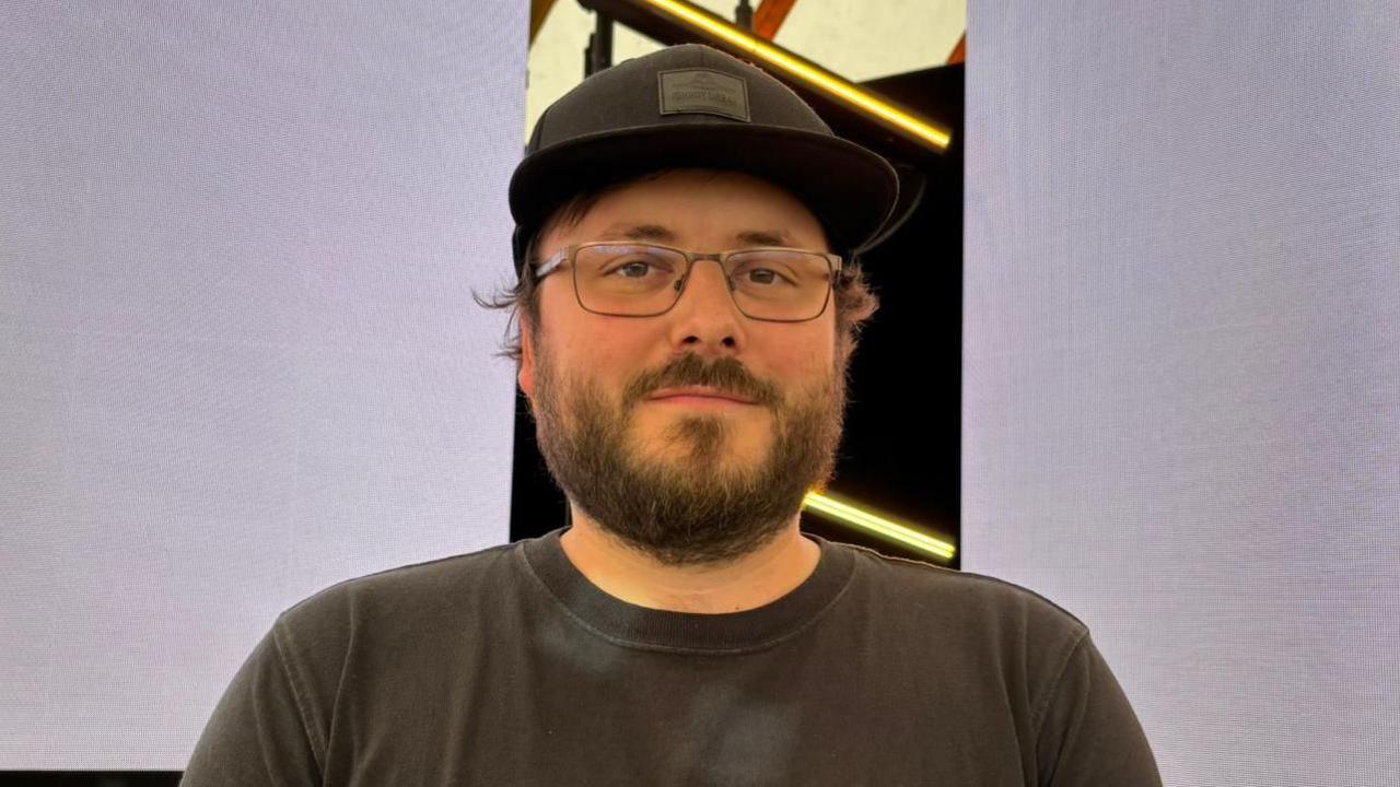 Joe Baker wearing a black sun cap and a black T-shirt and glasses. He has a black beard. He is smiling at the camera.