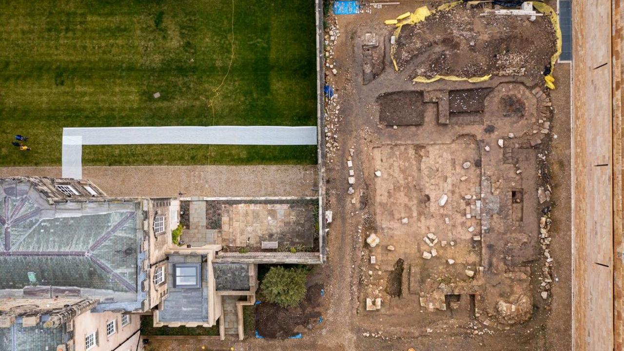 An aerial shot of the ruins. The land is dug and there are stones strewn across the site. There is a grass field to the left.