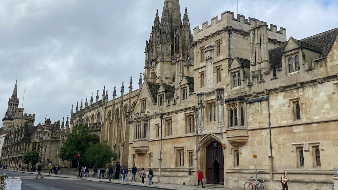 An historic building in Oxford with a number of spires. There are people walking on the pavement past outside. The sky is grey.