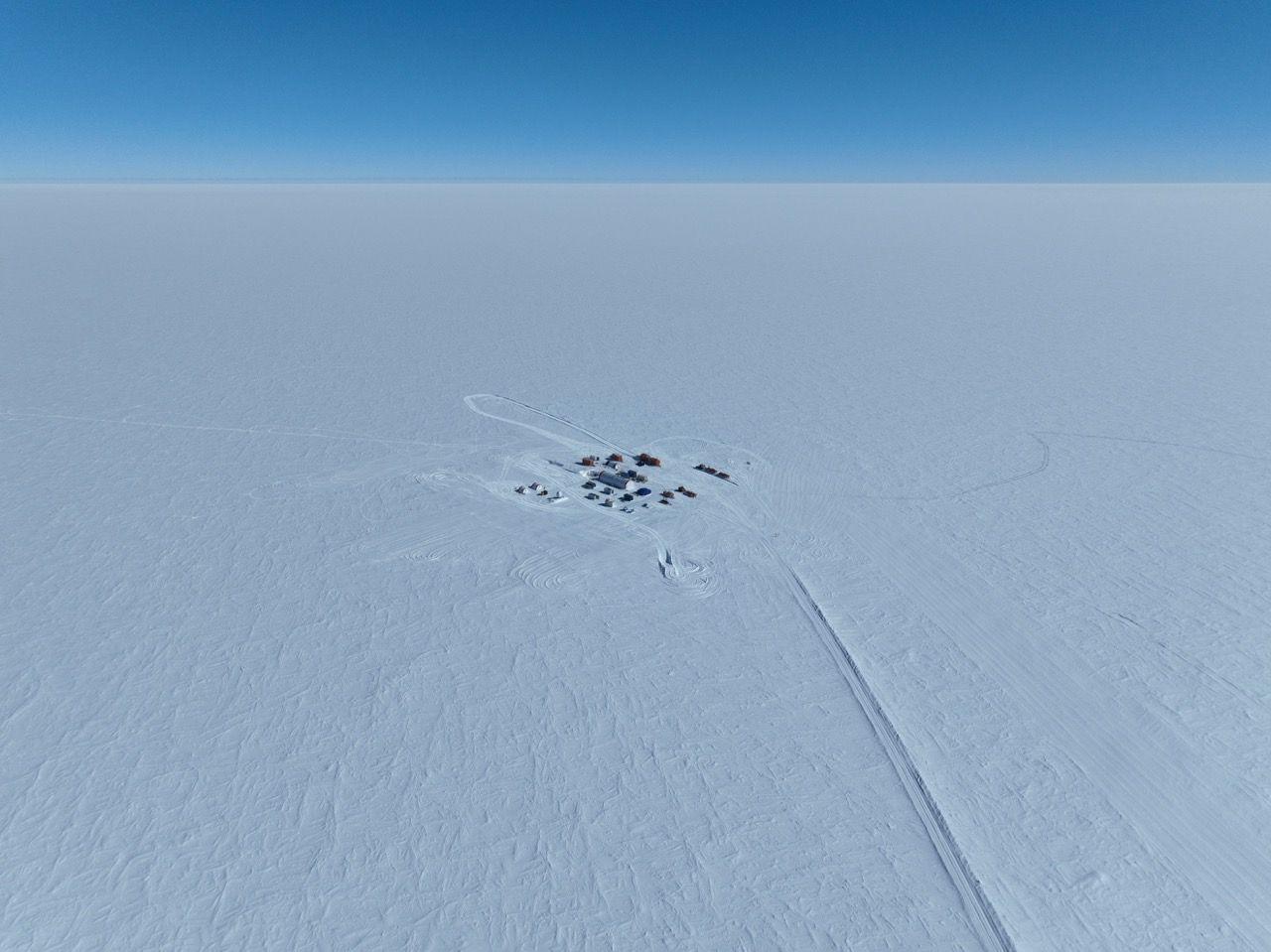A small group of tents and temporary shelters in the middle of the bright white Antarctic ice sheet. A trail of disturbed snow leads from the camp suggesting transport route. The sky is bright blue.